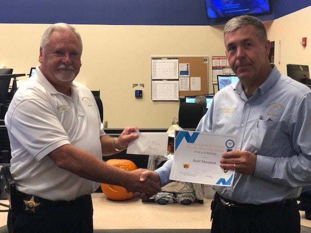 Two men shaking hands in an office, one holding a certificate and envelope.