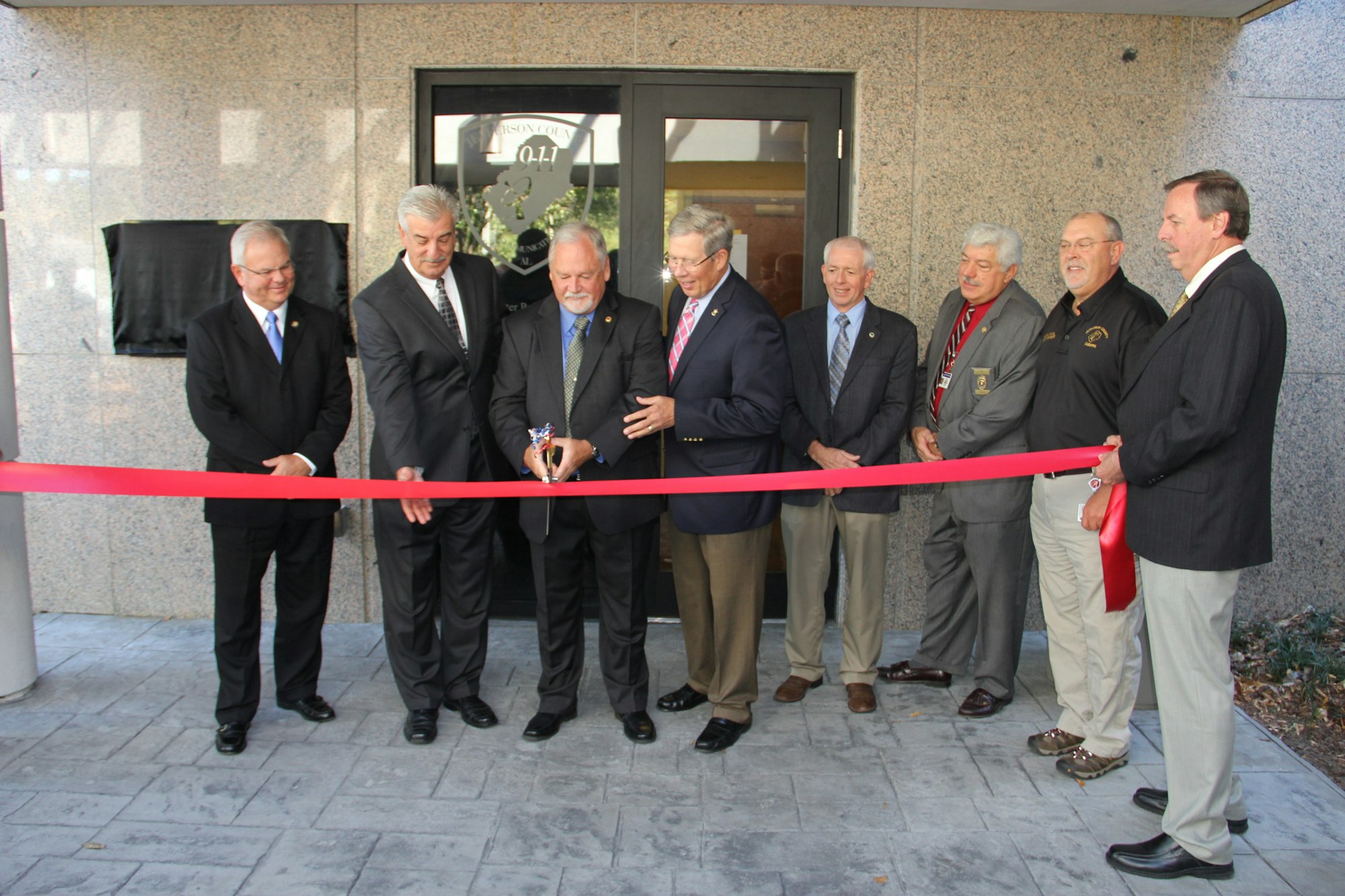 A group of men in suits cutting a red ribbon at an outdoor ceremony.