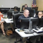 People working at desks with computers in an office setting, with a US flag in the background.