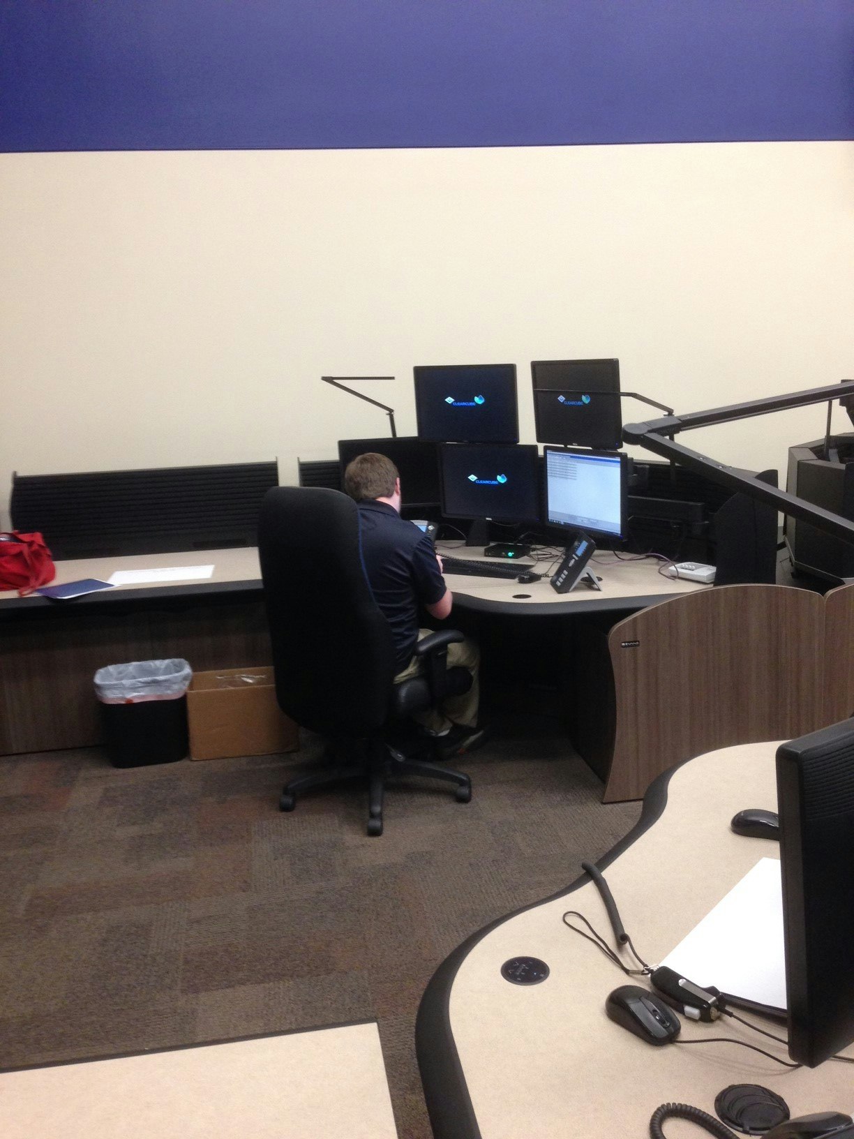 A person sits at a desk in a control room with multiple computer monitors and office equipment.