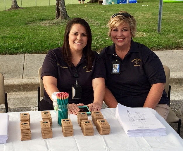 Two people sitting at a table with notepads and pencils, outdoors.