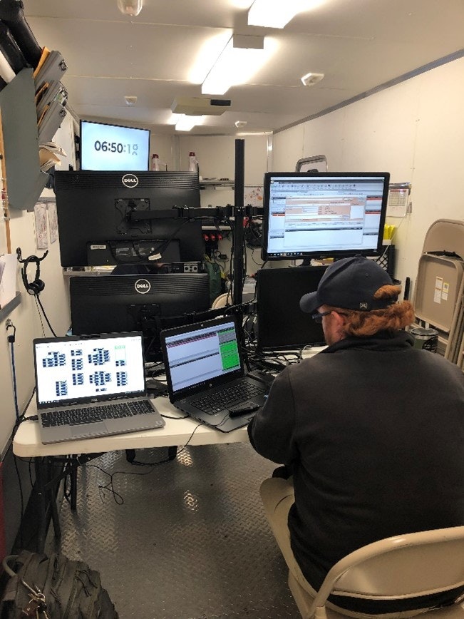 A person working at a station with multiple monitors displaying data and two laptops, in a well-lit room with a digital clock.
