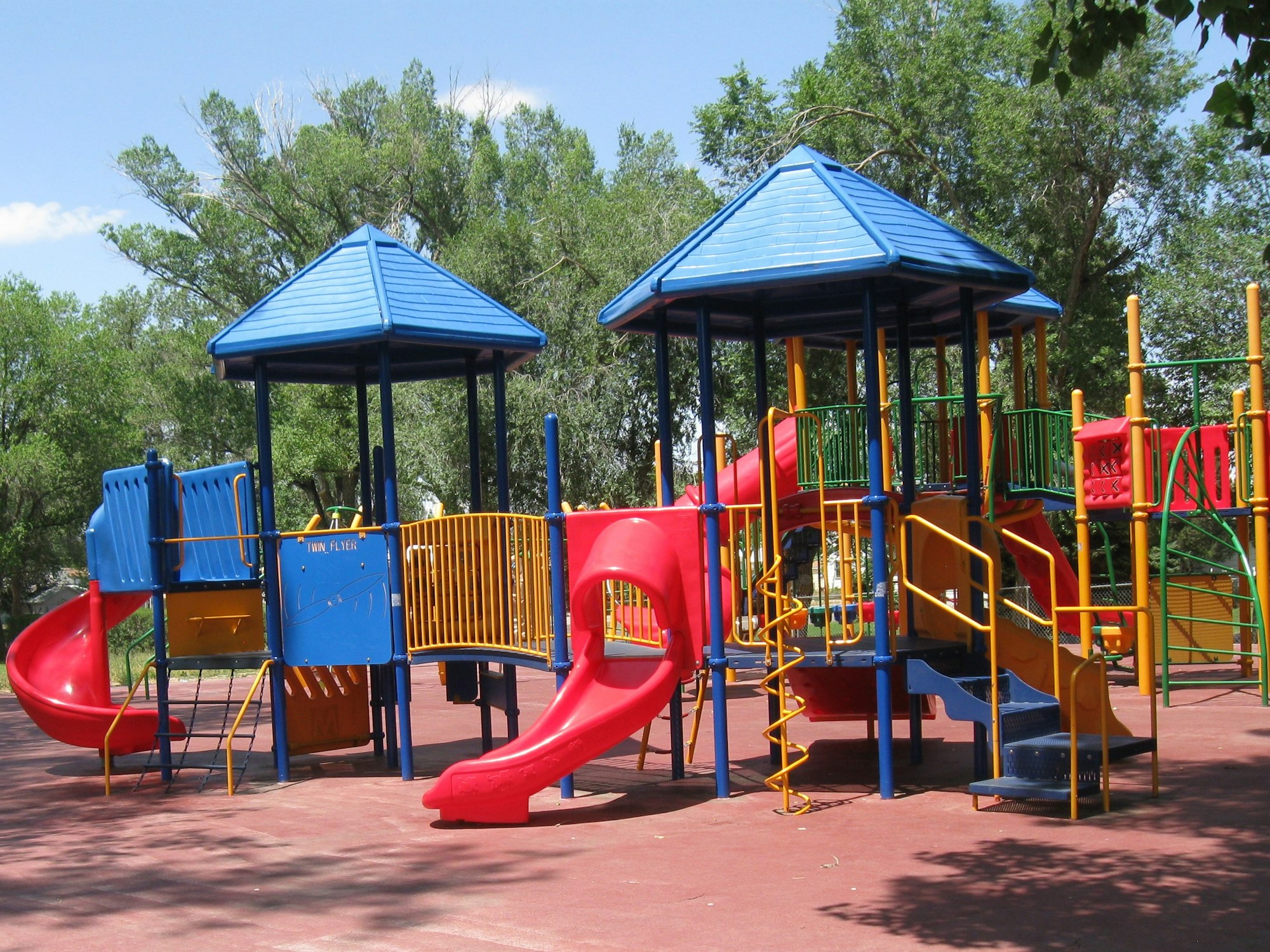 Colorful playground with slides and climbing structures, surrounded by trees and a clear blue sky.