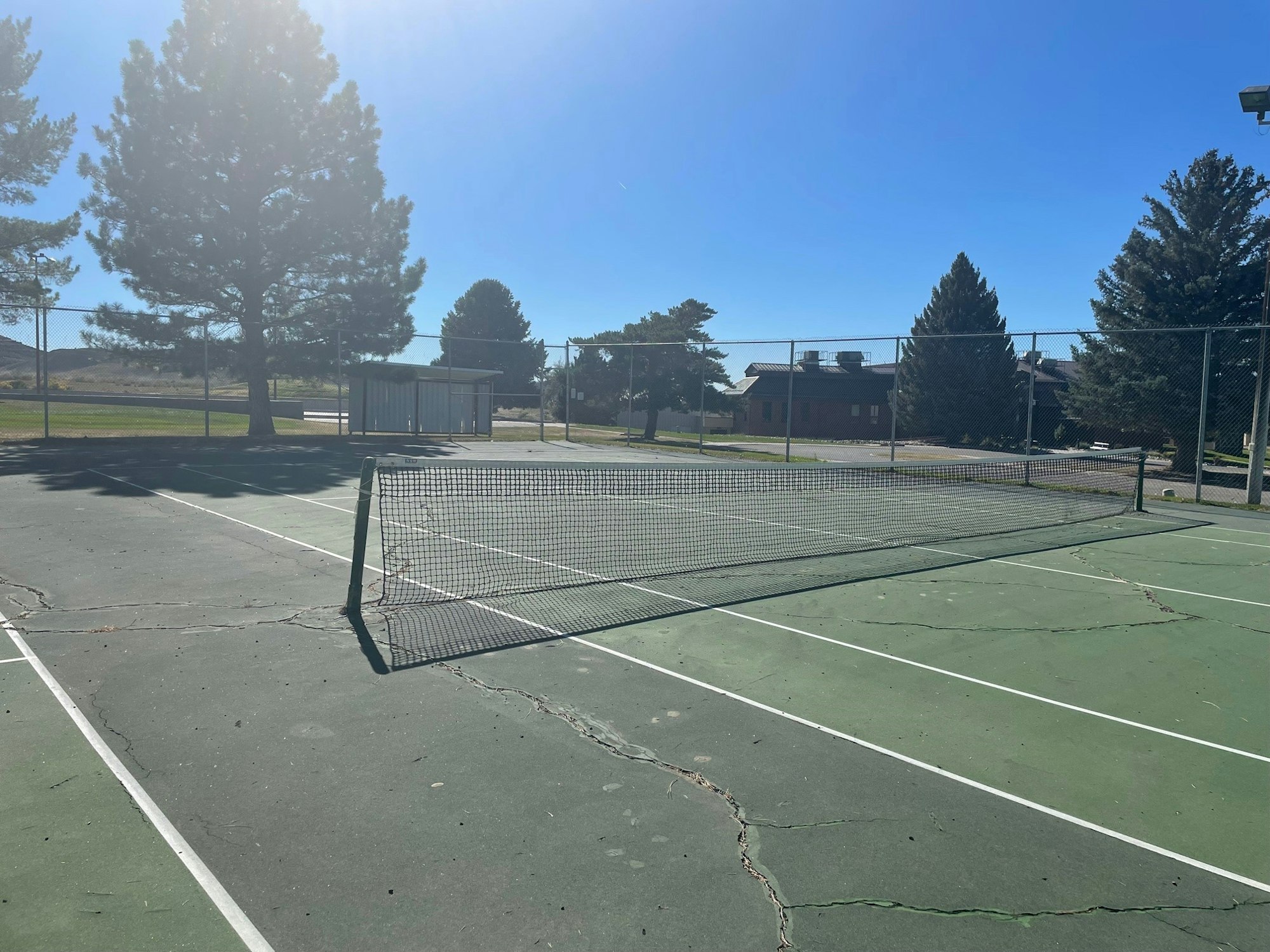 Outdoor tennis court with a net, surrounded by trees and buildings in the background.