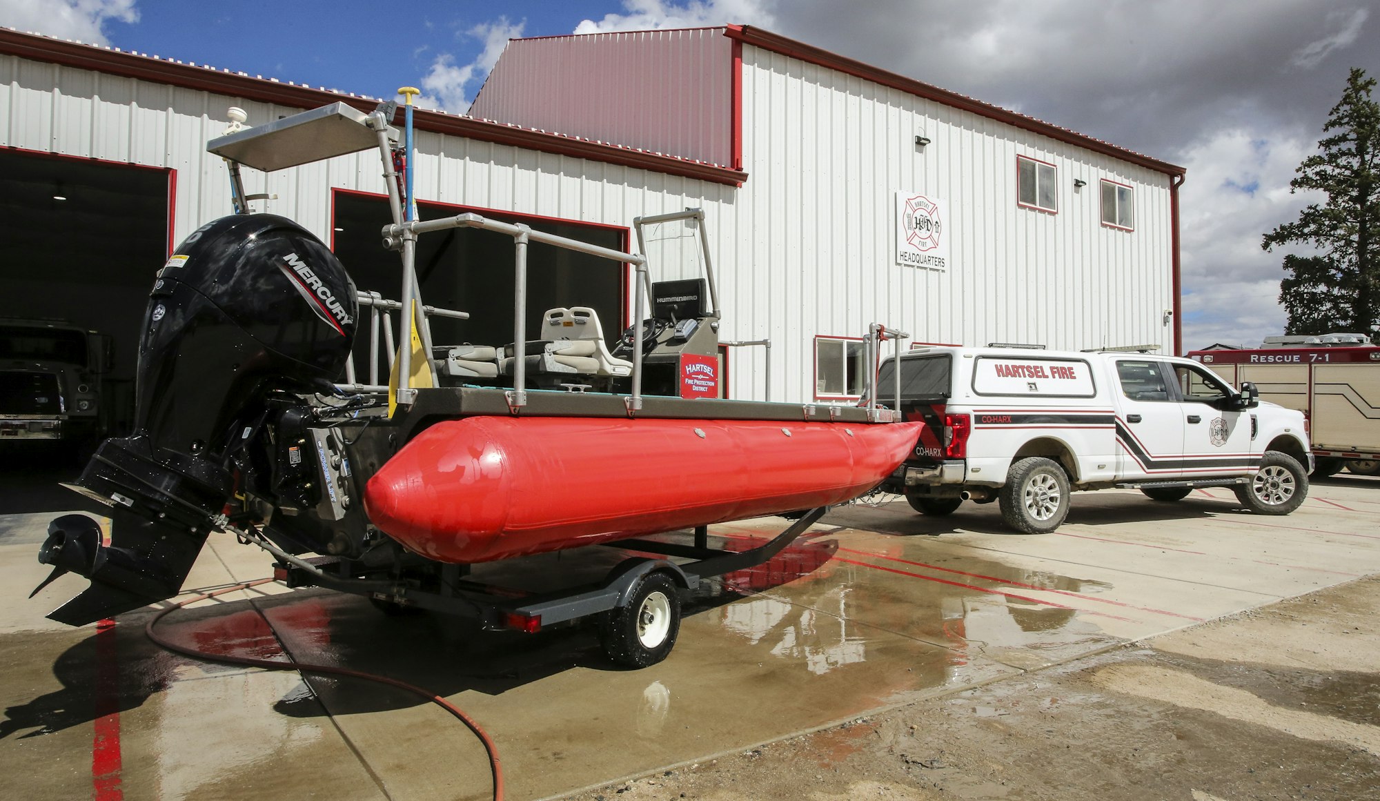 The image features a red rescue boat with a Mercury motor, parked outside the Hartsel Fire Department alongside a fire truck.