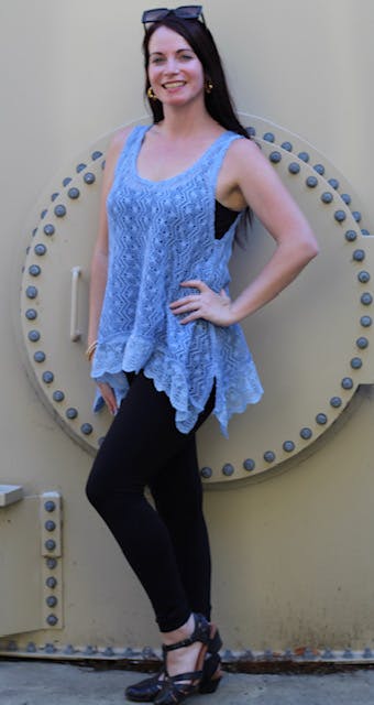 A person in a blue sleeveless top, black leggings, and sunglasses poses in front of a metal structure, smiling.