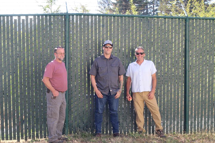 Three men standing in front of a tall green fence, one holding an apple, all wearing casual clothes and sunglasses in a sunny outdoor setting.