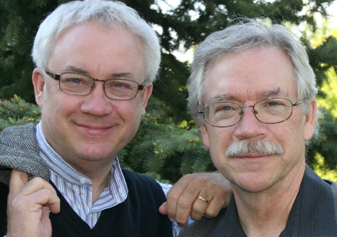 Two smiling men with glasses, one with his arm over the other's shoulder, standing outdoors with trees in the background.