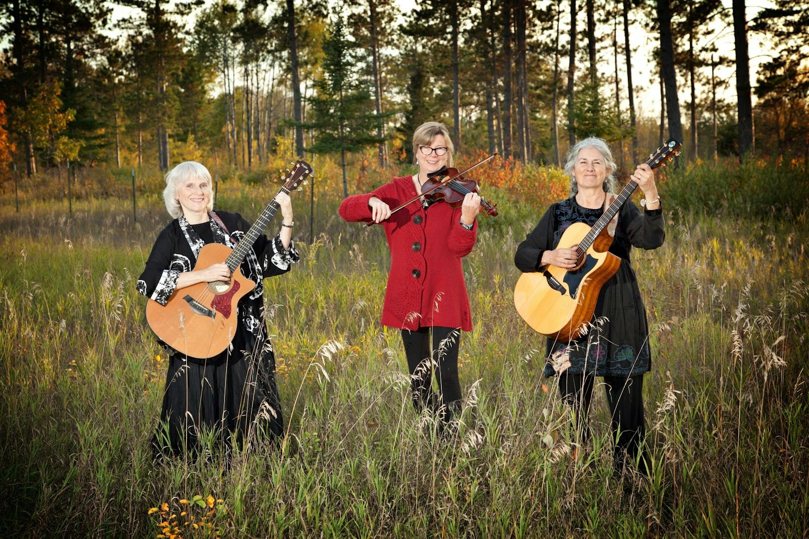 Three women in a field with two guitars and a violin, surrounded by trees.