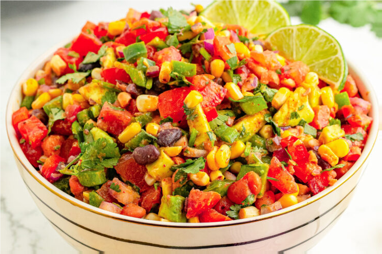 Colorful bowl of salsa with tomatoes, corn, beans, avocado, and cilantro, garnished with lime slices.