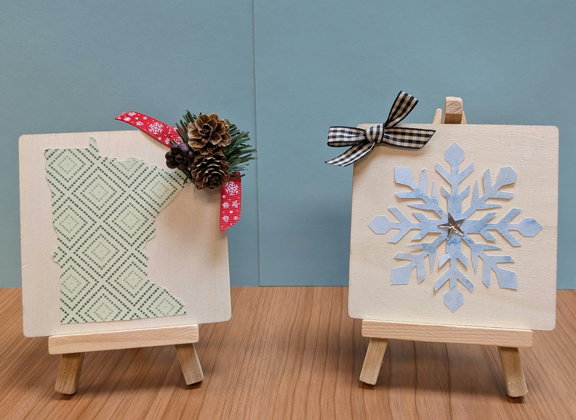 Two wooden art pieces on easels: one features a patterned outline of Minnesota with decorations, the other a blue snowflake design.