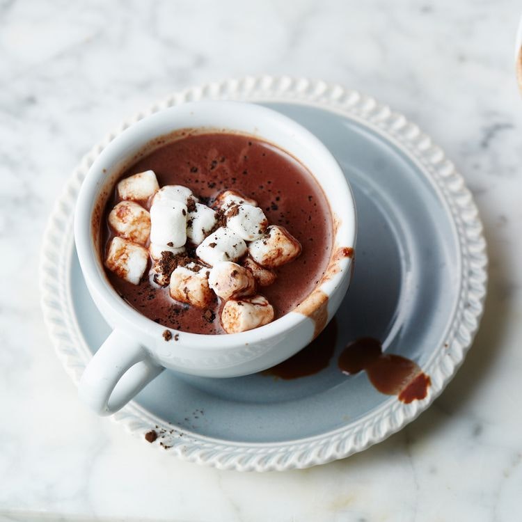 A cup of hot chocolate topped with marshmallows, sitting on a saucer, with some chocolate drips around.