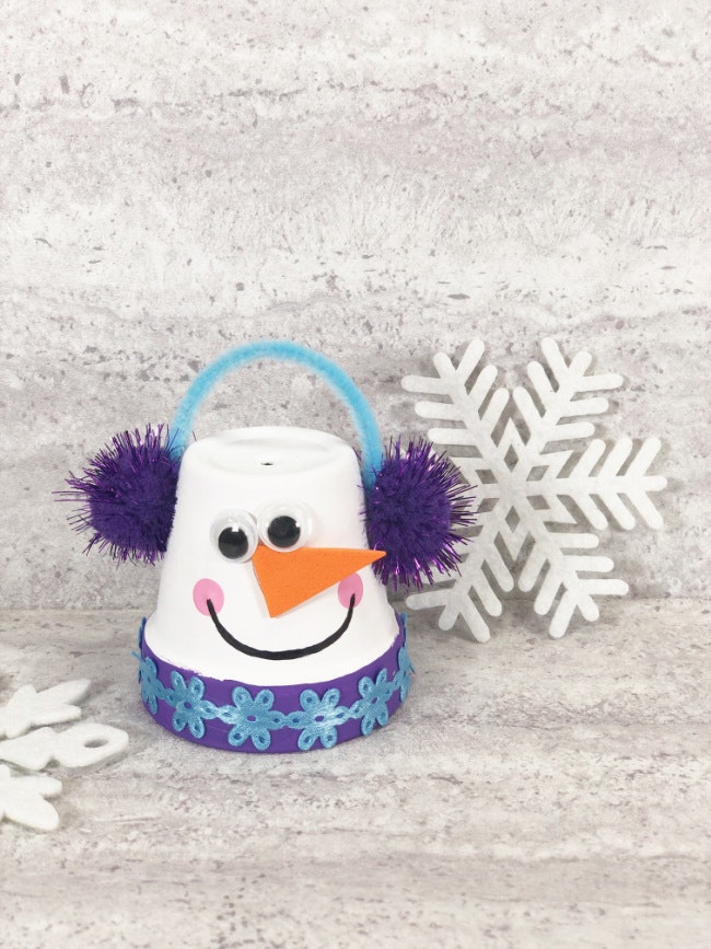 A craft snowman made from a cup with a blue handle, purple pom-poms, snowflakes, and a textured background.
