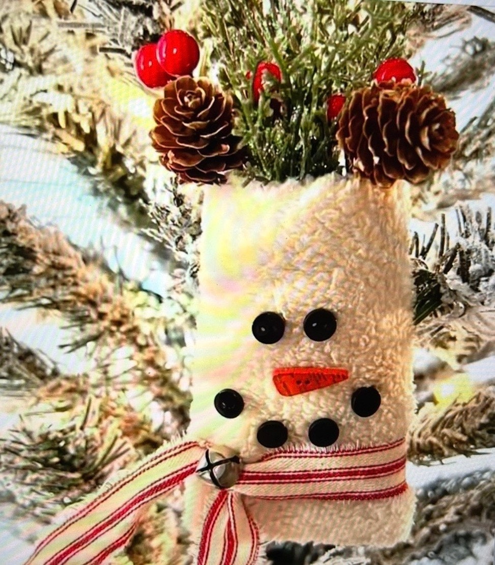 A cute, handcrafted snowman decoration made from fabric, adorned with buttons, pine cones, and red berries, hanging on a tree.