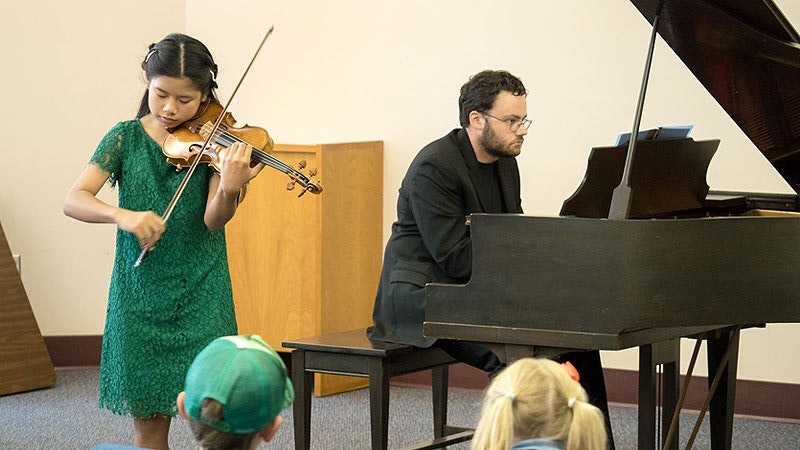 A girl in a green dress plays the violin while a man plays the piano; audience members are in the foreground.