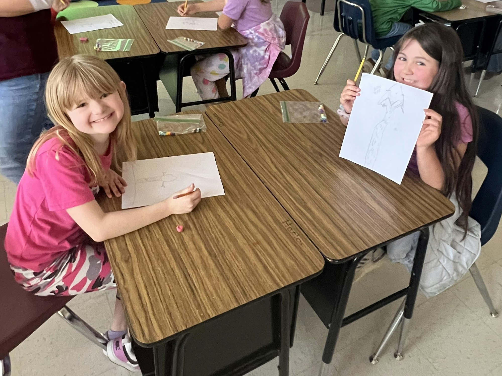 Two girls are smiling at a table in a classroom, one drawing while the other holds up her artwork.