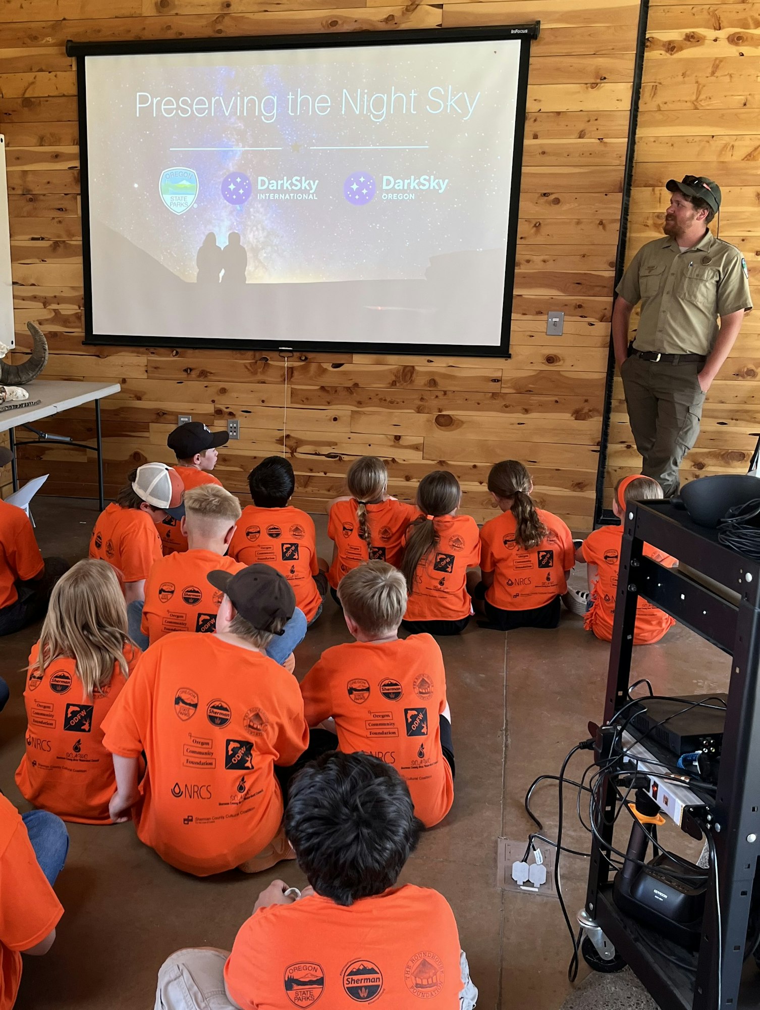A group of children in orange shirts watch a presentation titled "Preserving the Night Sky" in a wooden room.