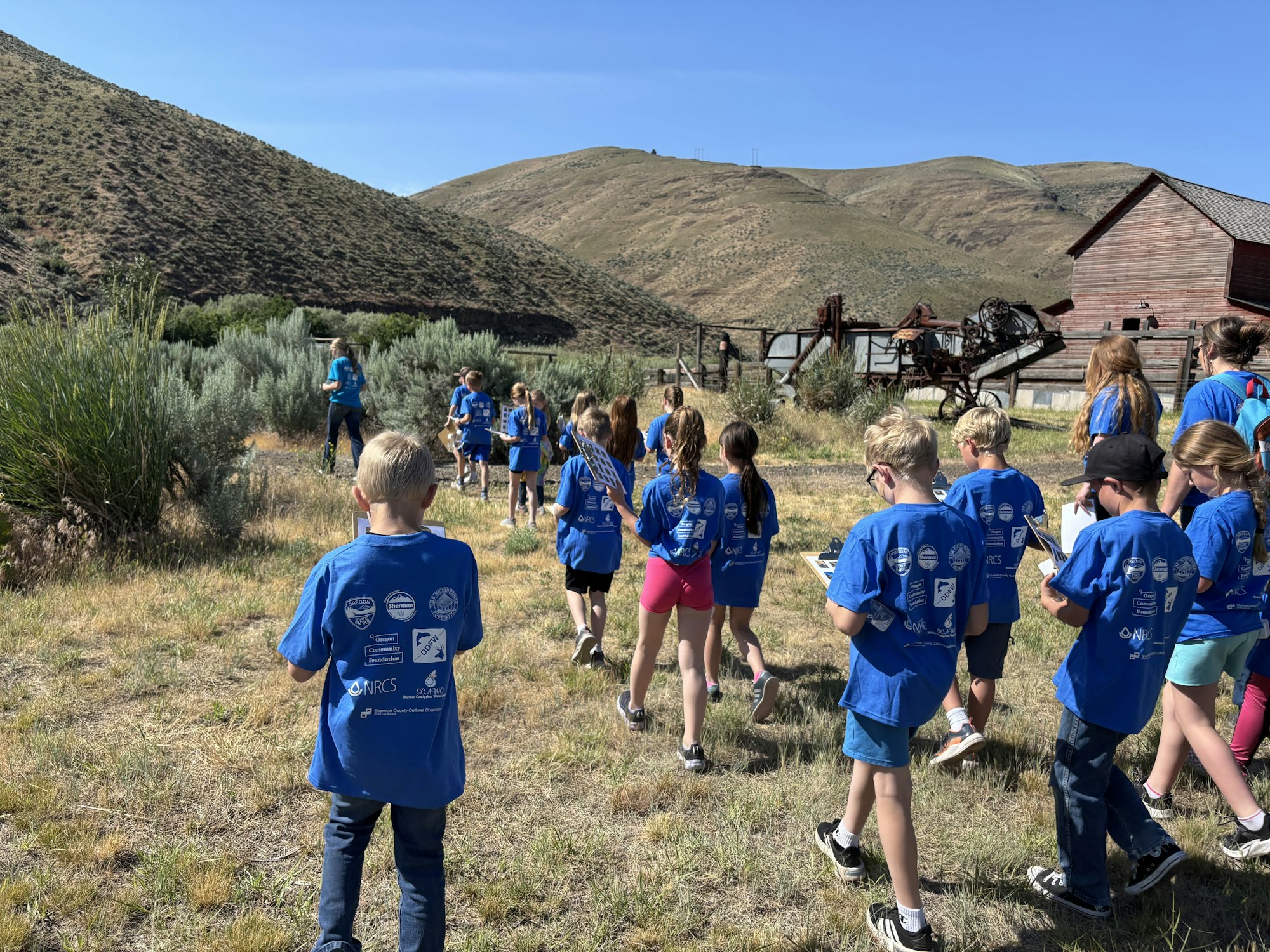 A group of children wearing blue shirts are walking outdoors near a wooden structure and hills.