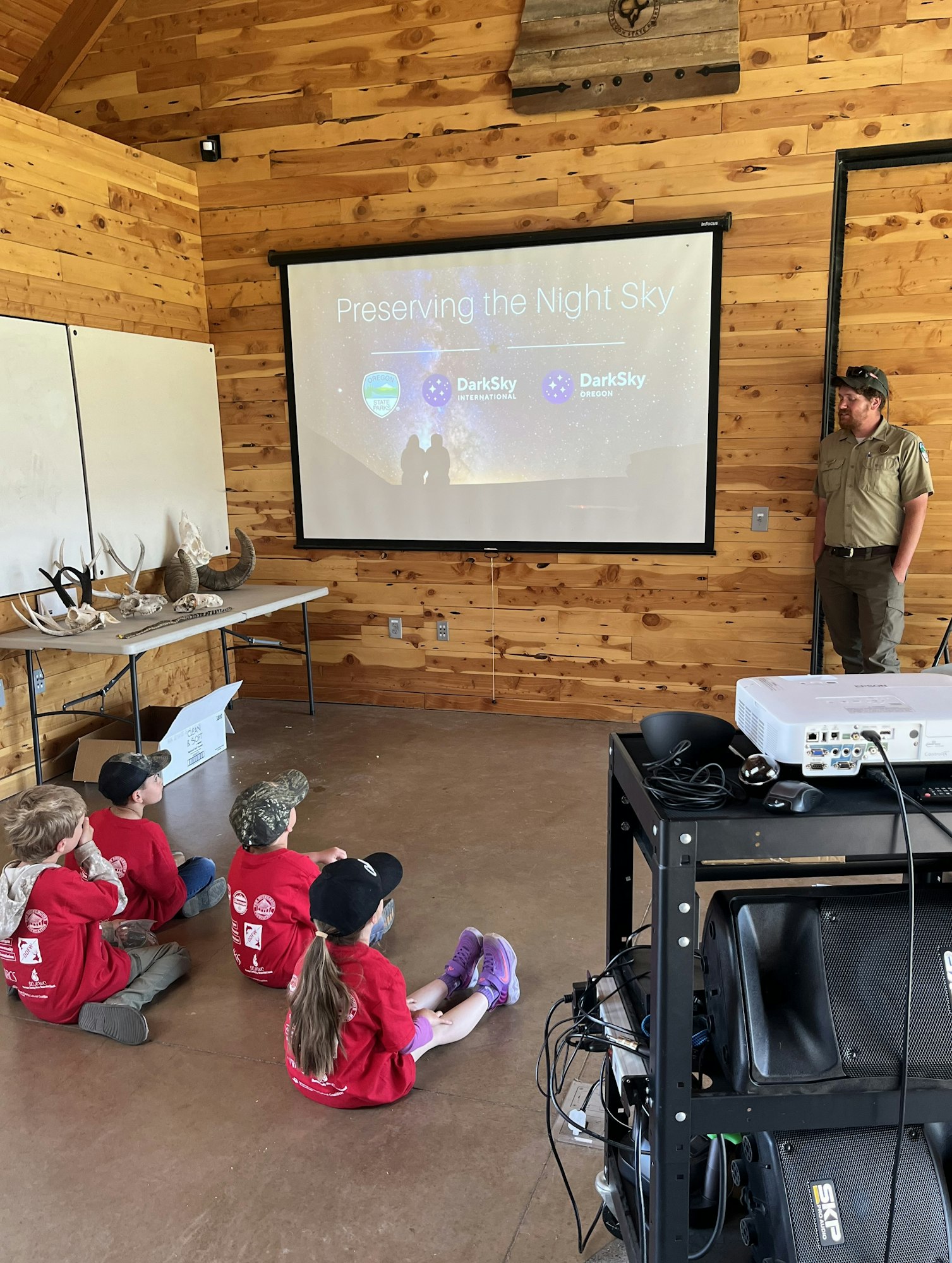 A presentation on "Preserving the Night Sky" to children in red shirts in a wooden room, featuring a projection screen and a speaker.