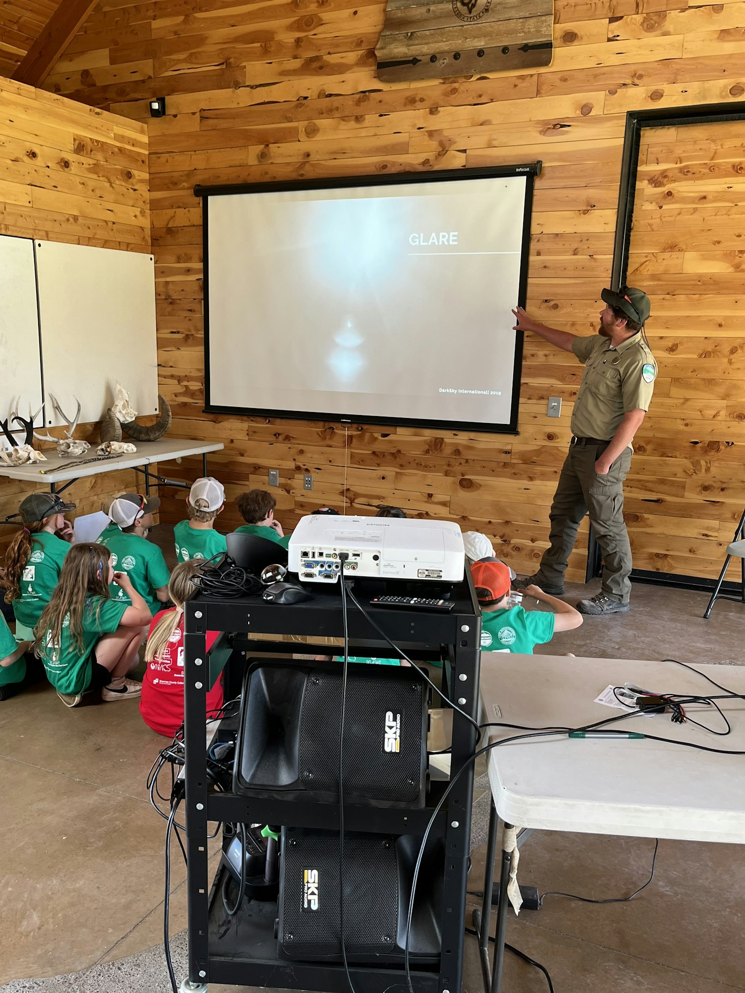 A person presents to children in a wooden room using a projector and screen displaying "GLARE."