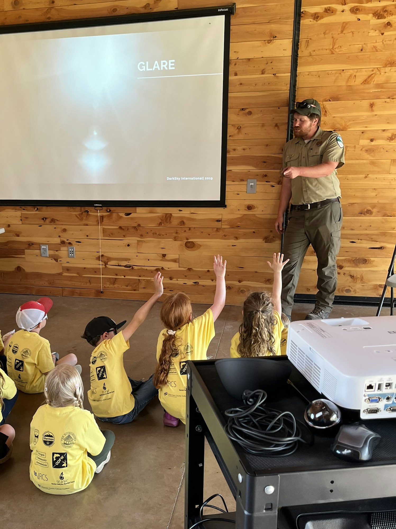 A group of children in yellow shirts raises hands during a presentation on glare led by an instructor.