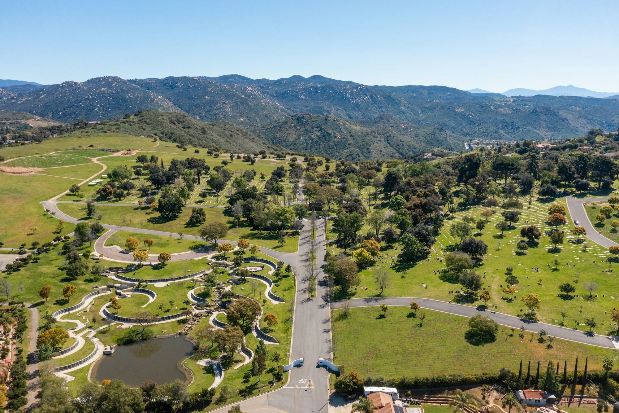 Aerial view of a landscaped cemetery with winding paths, a pond, and mountainous backdrop.