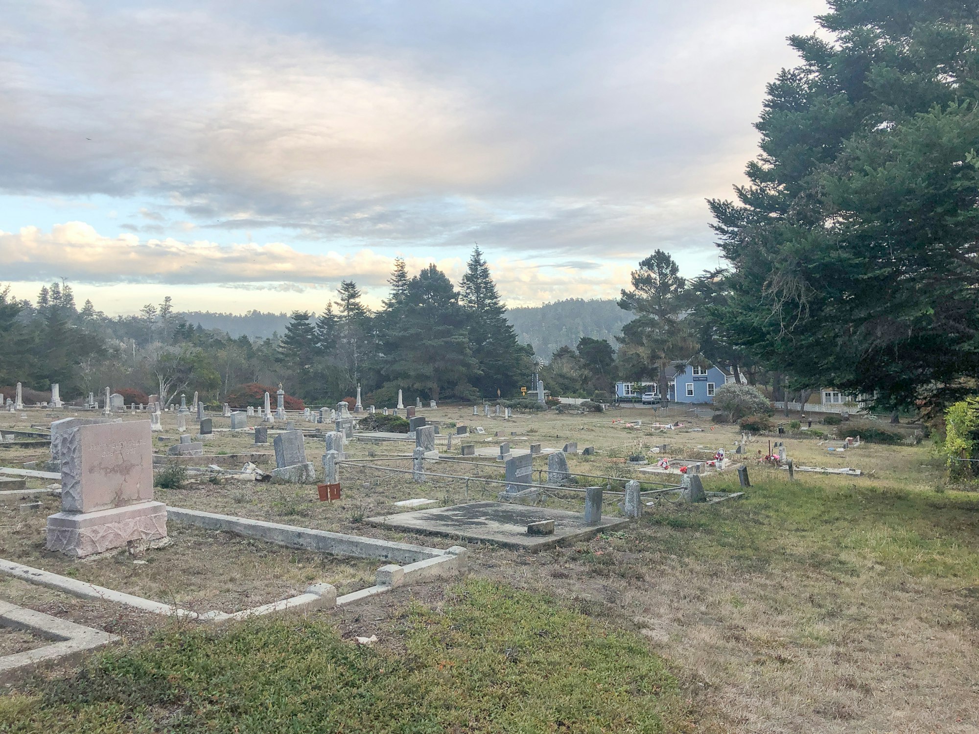 Photo of tombstones and a graveyard with beautiful clouds and a skyline created by trees.