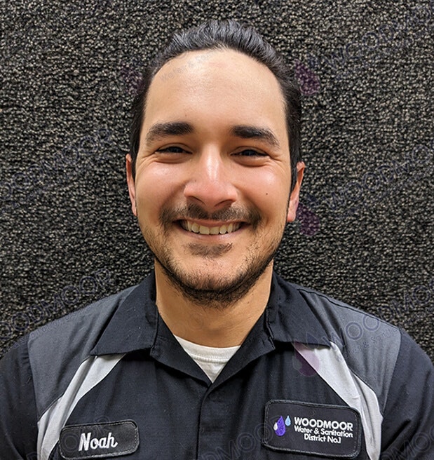 Smiling person in a uniform with name tag "Noah," in front of a textured background.