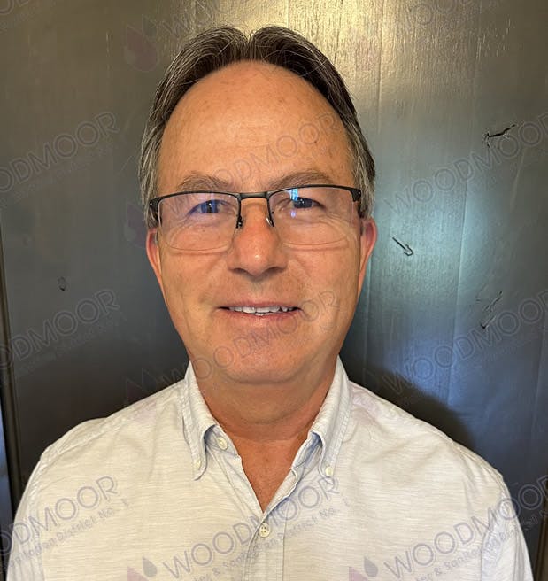 A person wearing glasses and a collared shirt, smiling at the camera against a plain background.