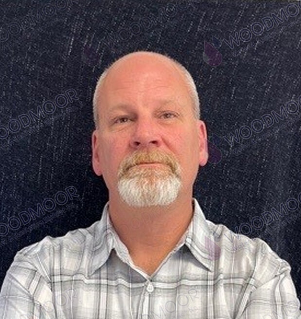 A man with a beard wearing a plaid shirt against a dark background with the watermark "WOODMOOR."