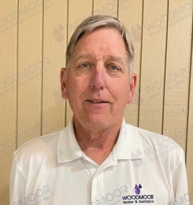 A man in a white shirt with "Woodmoor Water & Sanitation" logo stands against a wooden panel wall.