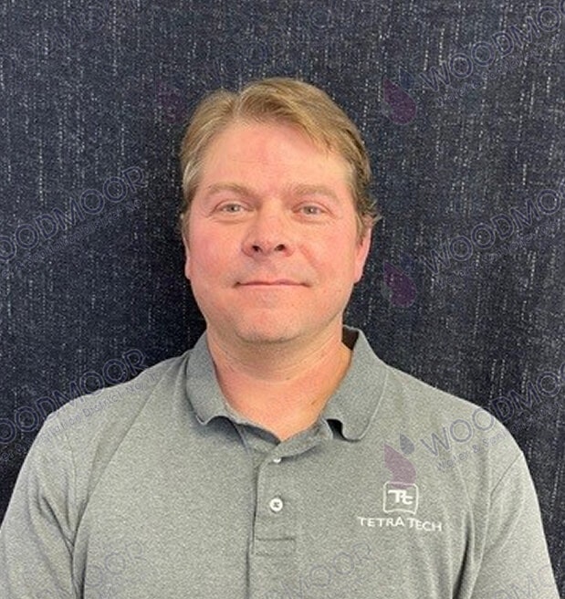 A person in a gray polo shirt with "Tetra Tech" logo, standing against a dark background with "WOODMOOR" watermark.