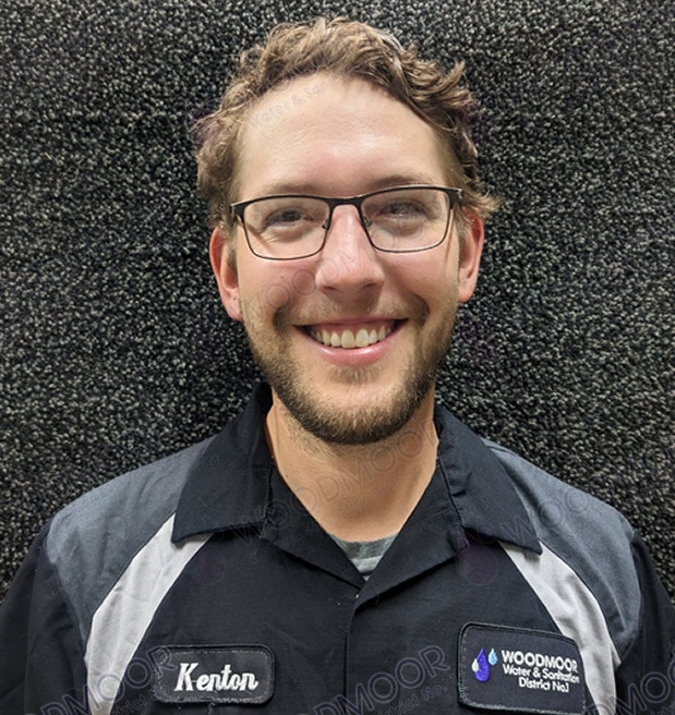 A smiling person wearing glasses and a shirt with name tags, standing against a textured background.