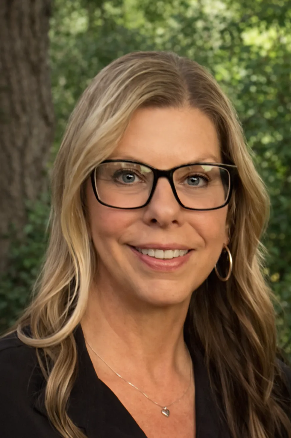 The image features a woman with long blonde hair, wearing glasses and a black top, smiling in a natural outdoor setting.