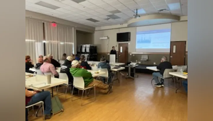 A group of people seated at tables in a meeting room, with a presenter speaking and a projector displaying data on a screen.