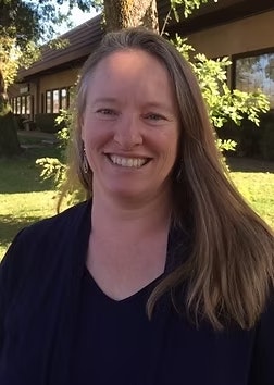 A person smiling outdoors, with trees and a building in the background.