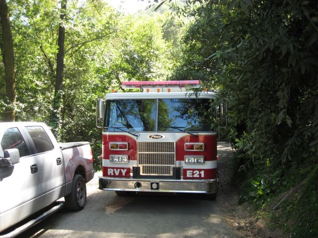 Red fire truck labeled "RVY E21" on a narrow, tree-lined road next to a parked white truck.