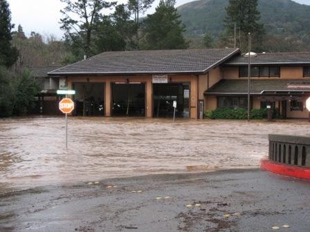 A building surrounded by floodwaters, with partially submerged road signs and trees in the background.