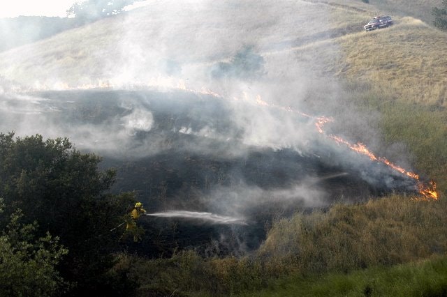 Firefighter extinguishing flames on a grassy hillside, with smoke rising and a vehicle on the slope.