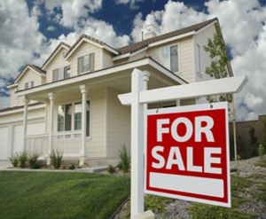 House with a "For Sale" sign in front, blue sky, and clouds in the background.