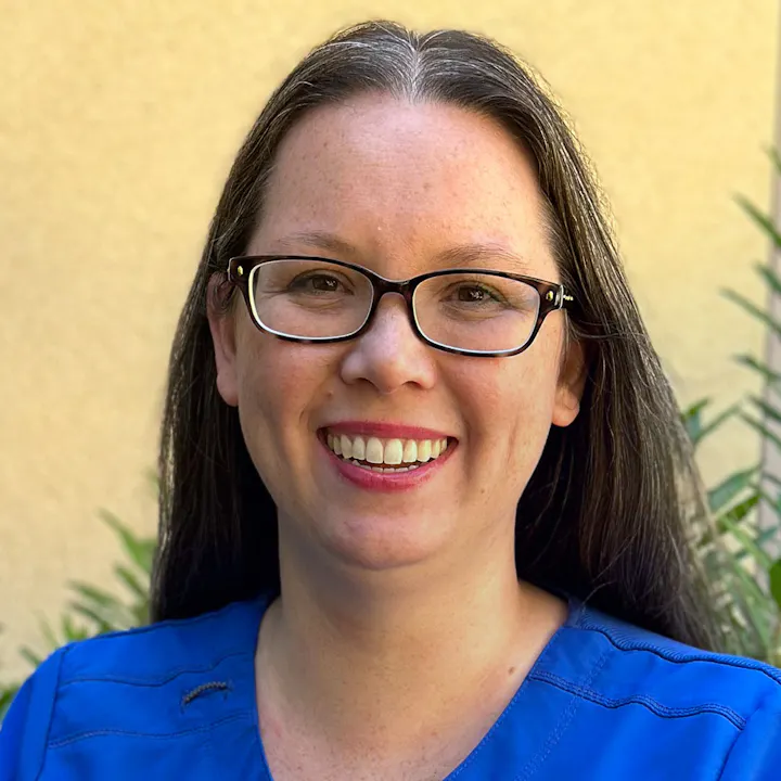 The image shows a smiling person with long hair, wearing glasses and a blue top, against a backdrop of greenery.