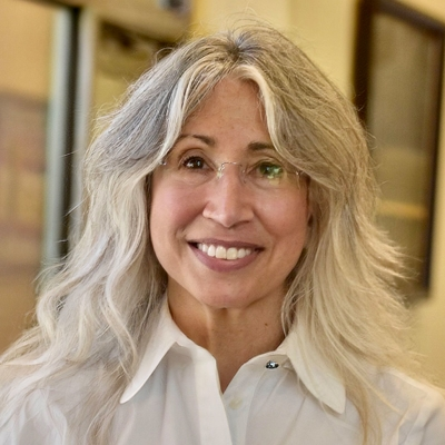 A smiling woman with long, wavy gray hair and glasses, wearing a white shirt, in a well-lit indoor setting.