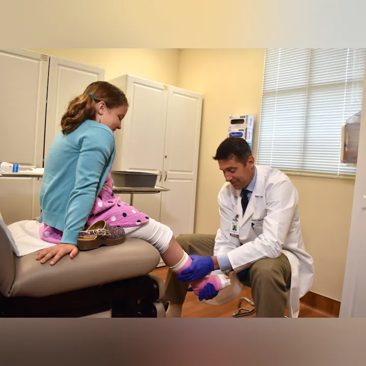 A doctor is examining a young girl's ankle in a healthcare setting. The girl appears relaxed while wearing a sweater and a blanket.