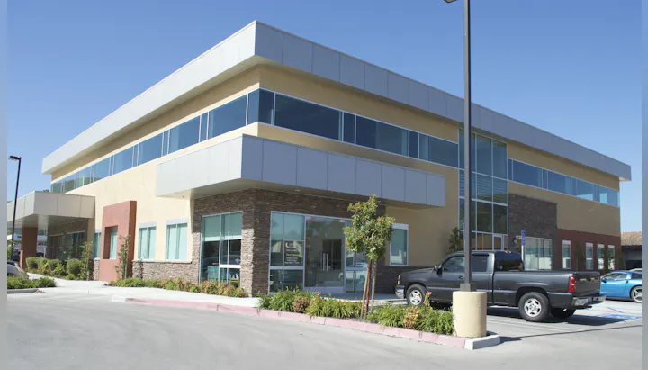 A modern commercial building with large windows, stone accents, landscaped surroundings, and parked vehicles.