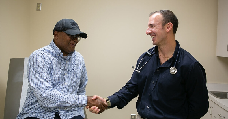 A patient and a doctor are shaking hands, smiling, in a medical office setting.