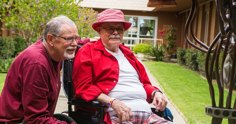 Two older men are enjoying time together outdoors, one in a wheelchair wearing a red shirt and hat, both smiling warmly.