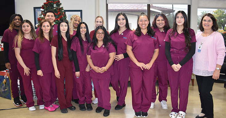 A group of healthcare professionals in maroon scrubs gathers for a photo, smiling in a festive environment with a holiday tree.