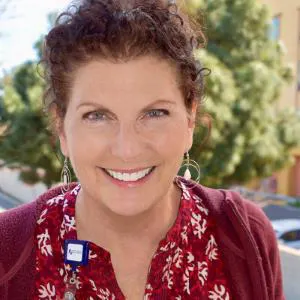 The image shows a smiling woman with curly hair, wearing a floral top and a cardigan, against a background of greenery.