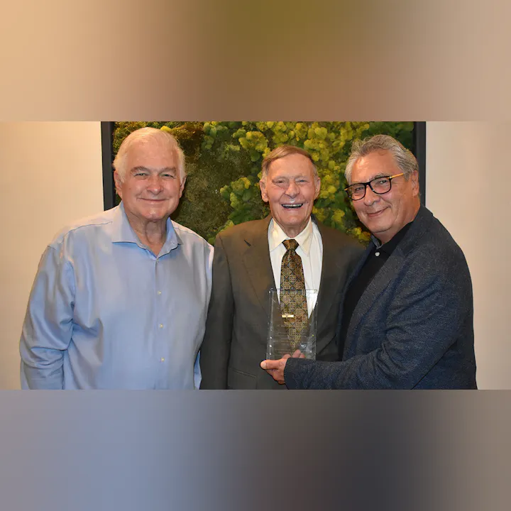 Three men pose together, smiling and holding a clear award, with greenery in the background. They appear to be celebrating an achievement.