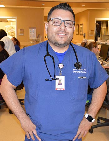 A smiling person in blue scrubs with a stethoscope around their neck, standing in a busy medical environment.